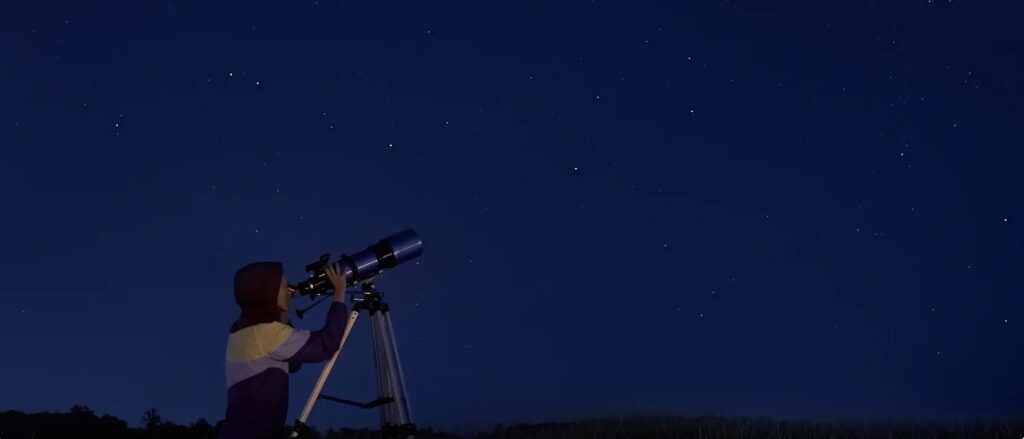 View of a person standing at a telescope looking toward stars in a dark blue night sky.