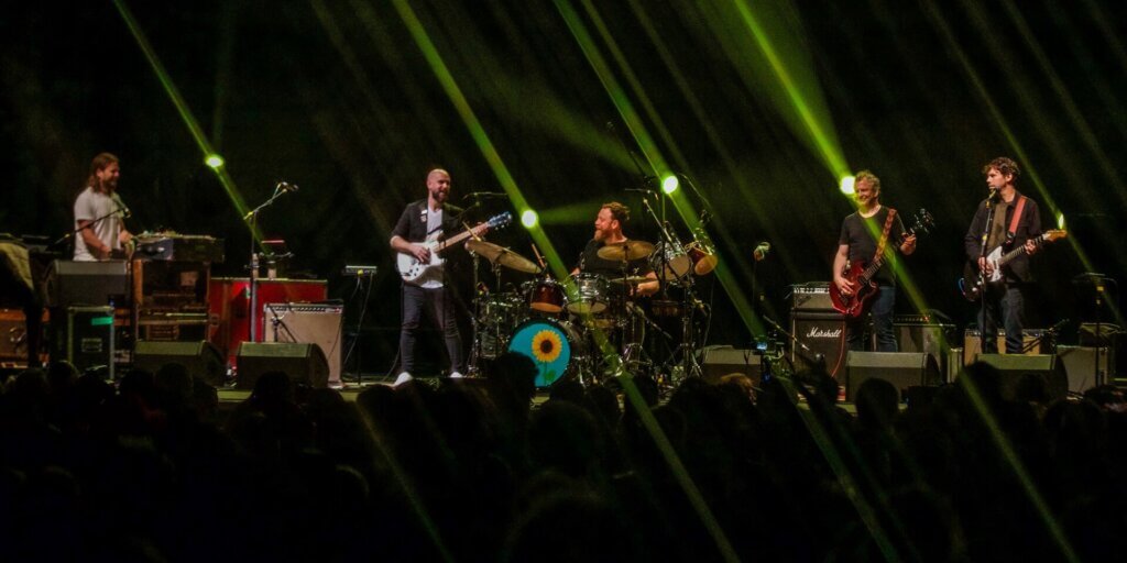 A five-piece band performs on a large, darkened concert stage. From left to right: a keyboardist at a keyboard rig surrounded by vintage amplifiers and equipment; a guitarist in a black blazer playing a white electric guitar; a bearded drummer seated behind a full kit, its kick drum painted with a teal and yellow sunflower; a guitarist playing a red electric guitar in front of a Marshall amp stack; and a second guitarist in a dark jacket playing an orange-tinted electric guitar. Green stage lights radiate diagonally across the scene. The silhouettes of a crowd are visible in the foreground.