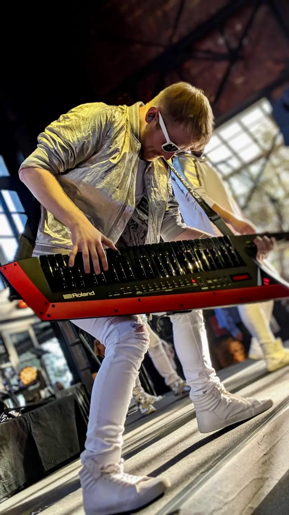 A dynamic, angled concert photograph of a keyboardist performing on an outdoor stage. He is bent forward intensely over a red Roland keytar, fingers pressing the keys as he plays. He wears white sunglasses, a shiny silver-gold jacket over a graphic t-shirt, white distressed jeans, and white high-top sneakers. An in-ear monitor is visible. The photo is shot from a low angle, giving the image a dramatic, energetic feel. Natural daylight streams through large windows or an open structure in the blurred background, with audience members partially visible.