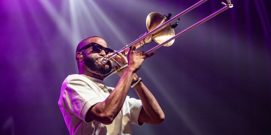 A close-up concert photograph of Trombone Shorty performing on stage under vivid purple and white spotlights. He is captured mid-performance, lips pressed to the mouthpiece of a gold and silver trombone, its slide extended upward and to the right. He wears dark wraparound sunglasses, an in-ear monitor, a cream short-sleeve shirt, a gold chain necklace, and a beaded bracelet on his right wrist. His expression conveys deep concentration and intensity. The background is a soft wash of purple light and haze.