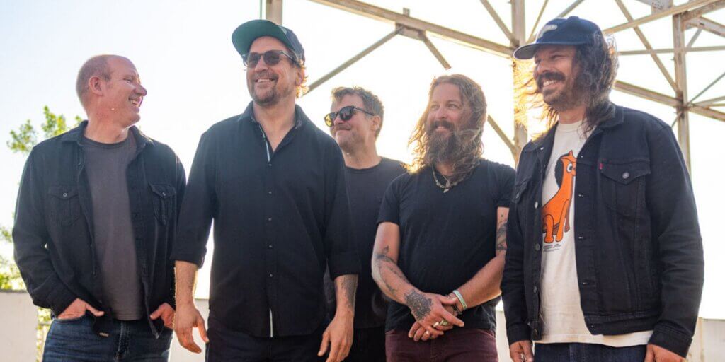 A candid photo of five adult men standing together outdoors in a row, smiling and laughing. From left to right: a man with short, buzzed hair in a dark button-up shirt; a taller man in a black button-up shirt and green baseball cap with sunglasses on his face; a man in a black t-shirt with yellow-tinted sunglasses; a man with long wavy hair and a full beard, tattoos on his forearms, wearing a black t-shirt and necklace; and a man with long curly dark hair in a white graphic t-shirt with an orange animal print layered under a dark denim jacket. The background shows a metal scaffolding structure and a bright, hazy sky with some foliage visible, suggesting an outdoor stage or festival setting. The mood is relaxed and joyful. Photo of the band Greensky Bluegrass.