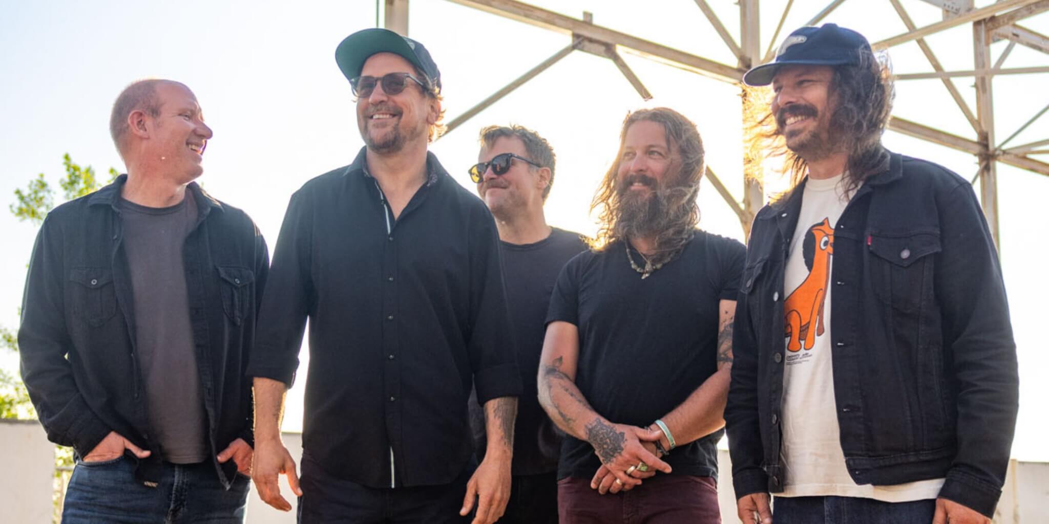 A candid photo of five adult men standing together outdoors in a row, smiling and laughing. From left to right: a man with short, buzzed hair in a dark button-up shirt; a taller man in a black button-up shirt and green baseball cap with sunglasses on his face; a man in a black t-shirt with yellow-tinted sunglasses; a man with long wavy hair and a full beard, tattoos on his forearms, wearing a black t-shirt and necklace; and a man with long curly dark hair in a white graphic t-shirt with an orange animal print layered under a dark denim jacket. The background shows a metal scaffolding structure and a bright, hazy sky with some foliage visible, suggesting an outdoor stage or festival setting. The mood is relaxed and joyful. Photo of the band Greensky Bluegrass.