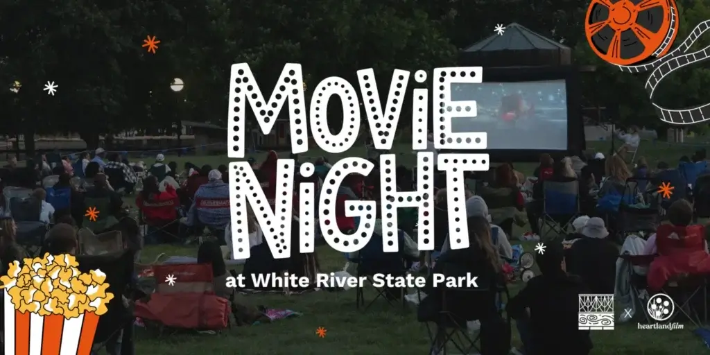 "Promotional graphic for 'Movie Night at White River State Park.' Background photo shows a crowd of people seated in lawn chairs and on blankets at dusk, watching a film on a large outdoor inflatable screen surrounded by trees. White polka-dot lettering reads 'MOVIE NIGHT' with 'at White River State Park' below it. An orange film reel with unspooling film strip decorates the top right corner, and an illustrated orange-and-white popcorn box sits in the bottom left corner, with white and orange sparkle/asterisk accents scattered throughout. Logos for White River State Park and Heartland Film appear in the bottom right.