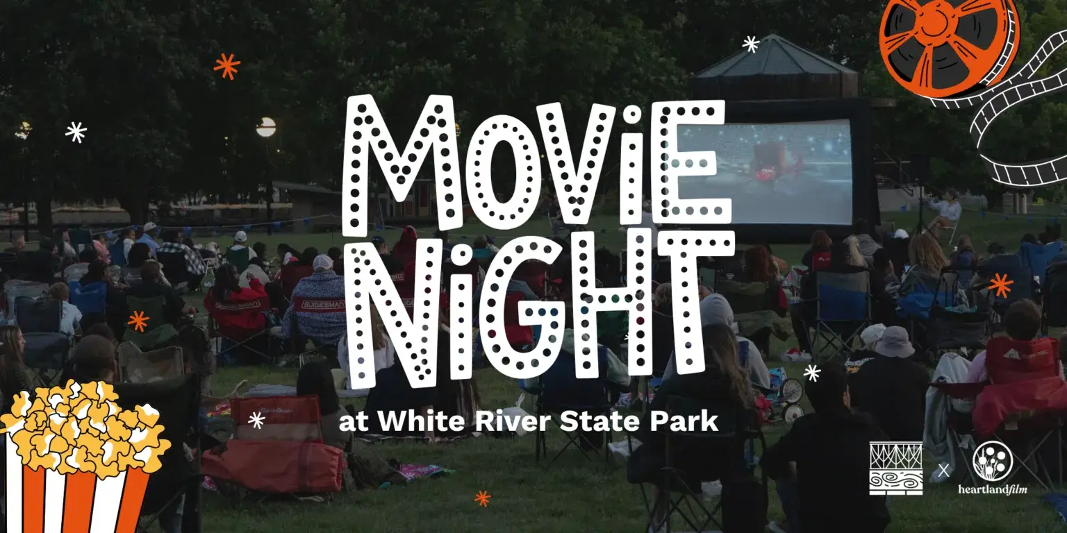 "Promotional graphic for 'Movie Night at White River State Park.' Background photo shows a crowd of people seated in lawn chairs and on blankets at dusk, watching a film on a large outdoor inflatable screen surrounded by trees. White polka-dot lettering reads 'MOVIE NIGHT' with 'at White River State Park' below it. An orange film reel with unspooling film strip decorates the top right corner, and an illustrated orange-and-white popcorn box sits in the bottom left corner, with white and orange sparkle/asterisk accents scattered throughout. Logos for White River State Park and Heartland Film appear in the bottom right.