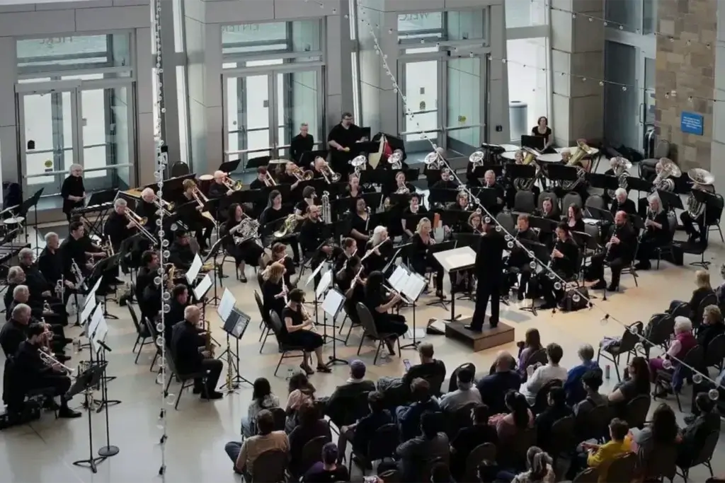 A wind ensemble performs in a bright, glass-walled atrium lobby, with musicians in black arranged in a semicircle around a conductor on a small podium. Rows of seated audience members watch from the foreground, and string lights are draped overhead near tall glass entrance doors.