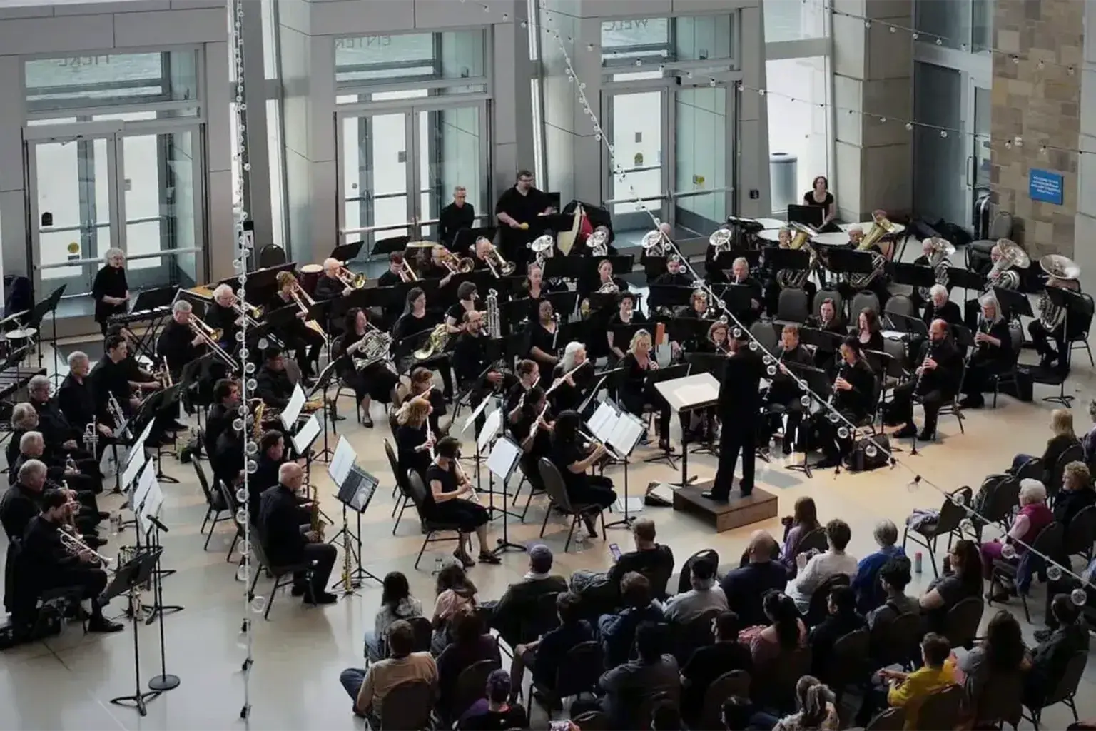 A wind ensemble performs in a bright, glass-walled atrium lobby, with musicians in black arranged in a semicircle around a conductor on a small podium. Rows of seated audience members watch from the foreground, and string lights are draped overhead near tall glass entrance doors.