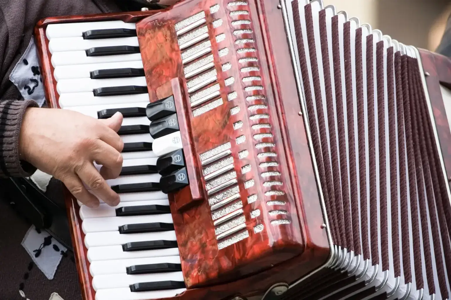 Close-up of a person's hand playing the keyboard of a red pearloid accordion, with the register switches, grille, and dark maroon bellows visible in the background.